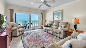 A living room with a ceiling fan, a sofa, a coffee table, a TV, and a view of the ocean through a sliding glass door. - Direct Beachfront Condo with Amazing Gulf Views! Pool, Grill, Free Wi-Fi & Parking! ~ 204 Seagate (Indian Rocks Beach)