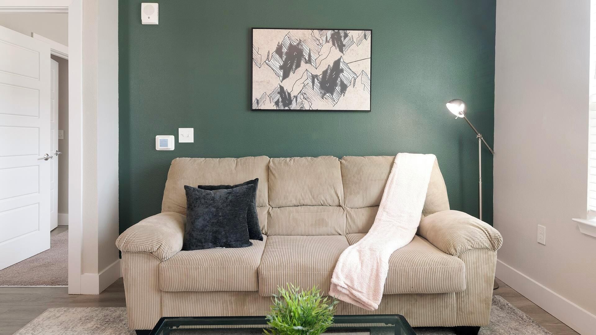 Una sala de estar con un sofá color beige, un cojín oscuro, una manta blanca, una obra de arte enmarcada, una lámpara de pie y una planta verde pequeña sobre una mesa de centro.
