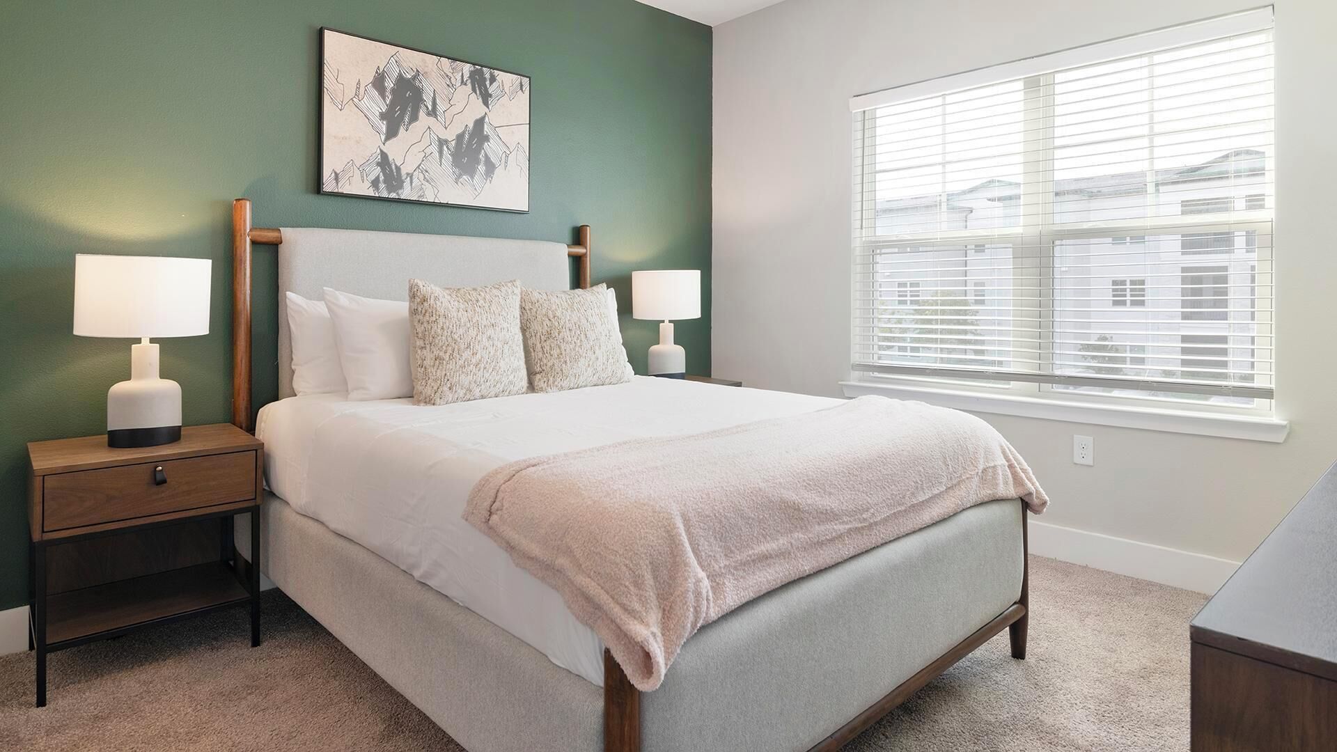 A bedroom with a bed, two bedside tables, a window with blinds, and a wall-mounted artwork.