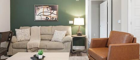 Un salón con un sofá color beige, un sillón de cuero marrón, una mesa de centro con una planta y un control remoto, una lámpara y un cuadro en la pared.