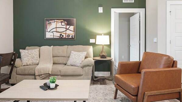 A living room with a beige sofa, a brown leather armchair, a coffee table with a plant and remote, a lamp, and a painting on the wall. - Landing | Modern 2BD, Clubhouse, Pool (Orlando)