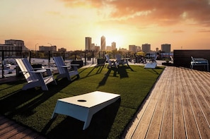 Eine Dachterrasse mit Liegestühlen und einem kleinen Tisch mit Blick auf eine städtische Skyline bei Sonnenuntergang.