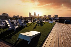 Eine Dachterrasse mit Liegestühlen und einem kleinen Tisch mit Blick auf eine städtische Skyline bei Sonnenuntergang.