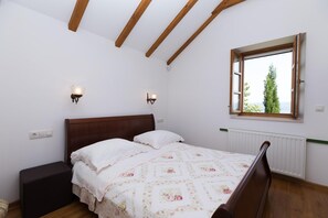 Ein Schlafzimmer mit Holzboden, einem Bett mit floralem Überwurf, zwei Wandleuchten und einem Fenster mit Blick auf Grünflächen.