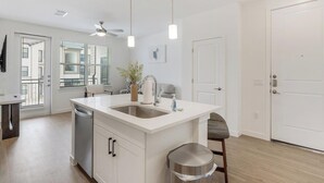 A modern kitchen with a central island, a dishwasher, and a sink. There is a ceiling fan, a window with blinds, and a door. - Landing | Stylish 1BD, Pool, Clubhouse (Dallas)