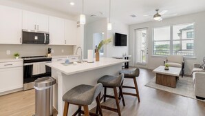 A modern kitchen with white cabinetry, a central island, bar stools, a stainless steel trash can, and a ceiling fan. - Landing | Stylish 1BD, Pool, Clubhouse (Dallas)