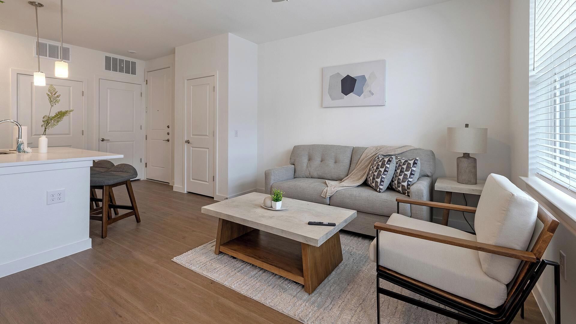 A modern living room with a sofa, coffee table, and chairs, a kitchen area with a sink and cabinets, and a window with blinds.