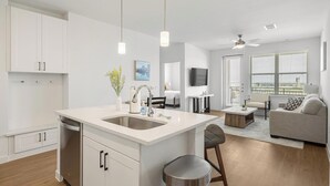 A modern kitchen with a central island, a dining area with a sofa and coffee table, and a living room with a TV and seating. - Landing | Modern 2BD, Clubhouse, Courtyard (Dallas)