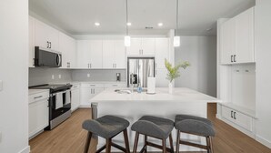 A modern kitchen with white cabinets, a central island, bar stools, and a microwave. - Landing | Modern 2BD, Clubhouse, Courtyard (Dallas)