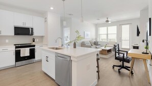 Cozinha moderna com lava-louças, fogão e micro-ondas. Há uma sala de jantar com mesa e cadeiras, e uma sala de estar com sofá e mesa de centro.