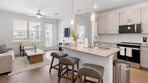 A modern kitchen with a central island, a dining area with a sofa and chairs, and a living room with a TV and a plant. - Landing | Amazing 1BD, Gym, Clubhouse (Dallas)