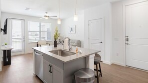 A modern kitchen with a central island, a ceiling fan, and a view of the outside through large windows. - Landing | Modern 1BD, Courtyard, Gym (Dallas)