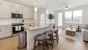 A modern kitchen with a central island, bar stools, a dining area with a table and chairs, and a living room with a sofa and a TV. - Landing | Beautiful 1BD, Gym, Clubhouse (Dallas)