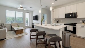 A modern kitchen with a central island, a dining area with a sofa and chairs, and a TV mounted on the wall. - Landing | Amazing 1BD, Courtyard, Clubhouse (Dallas)
