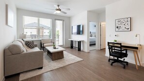 A modern living room with a sofa, coffee table, and a TV. There's a desk with a chair and a ceiling fan. - Landing | Chic 2BD, Courtyard, Pool (Dallas)