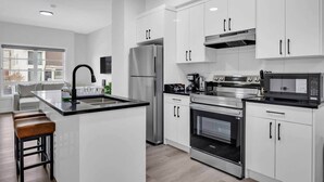 A modern kitchen with white cabinets, stainless steel appliances, a central island with stools, and a large window providing a view of the city. - 105-Urban Retreat / Stylish-brand-new modern home (Calgary)