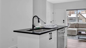 A modern kitchen with a black countertop, a built-in dishwasher, and a window overlooking a residential area. - 105-Urban Retreat / Stylish-brand-new modern home (Calgary)