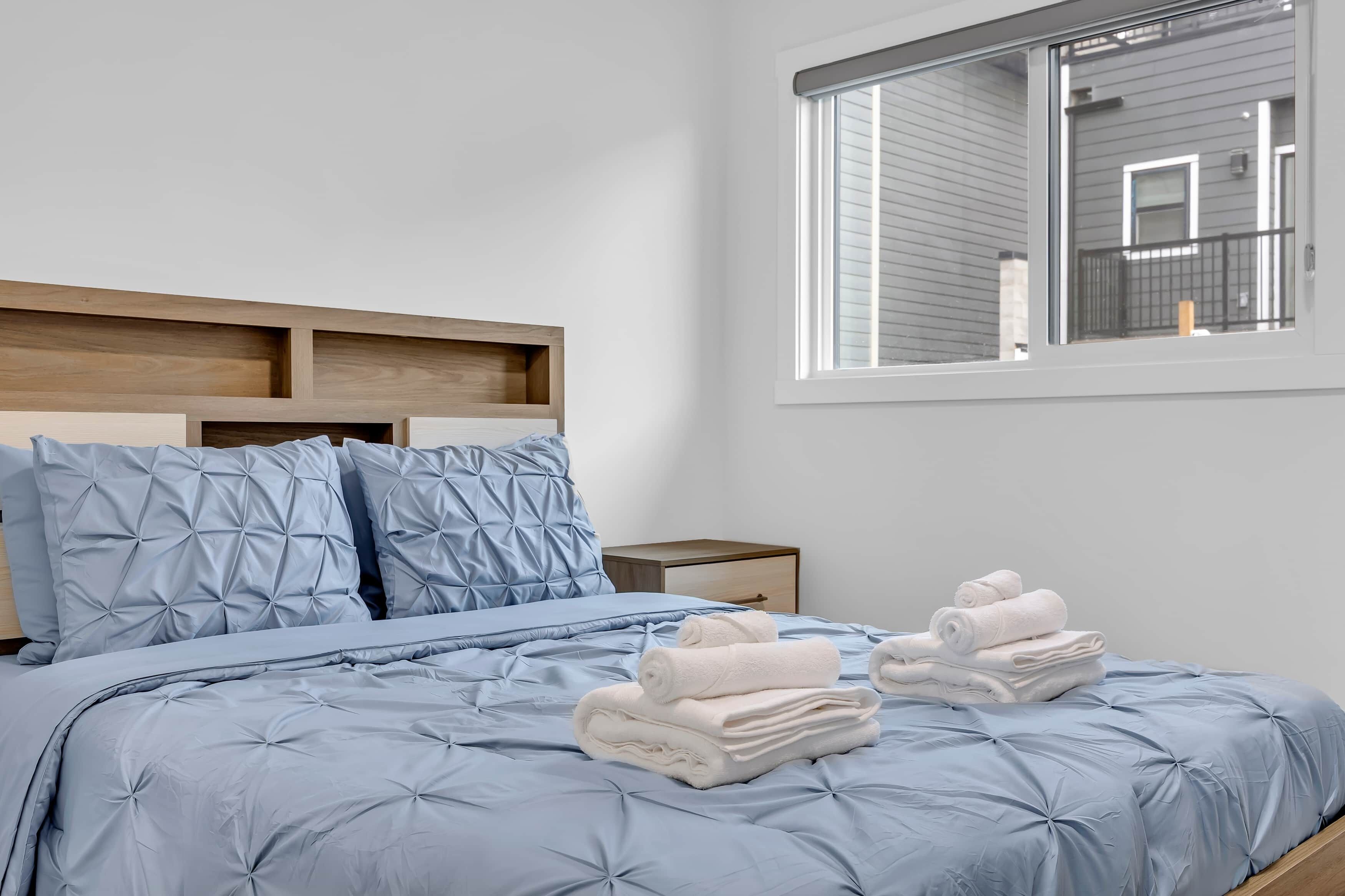 A neatly made bed with a blue quilt, pillows, and rolled towels.