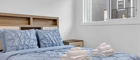 A neatly made bed with a blue quilt, pillows, and rolled towels.