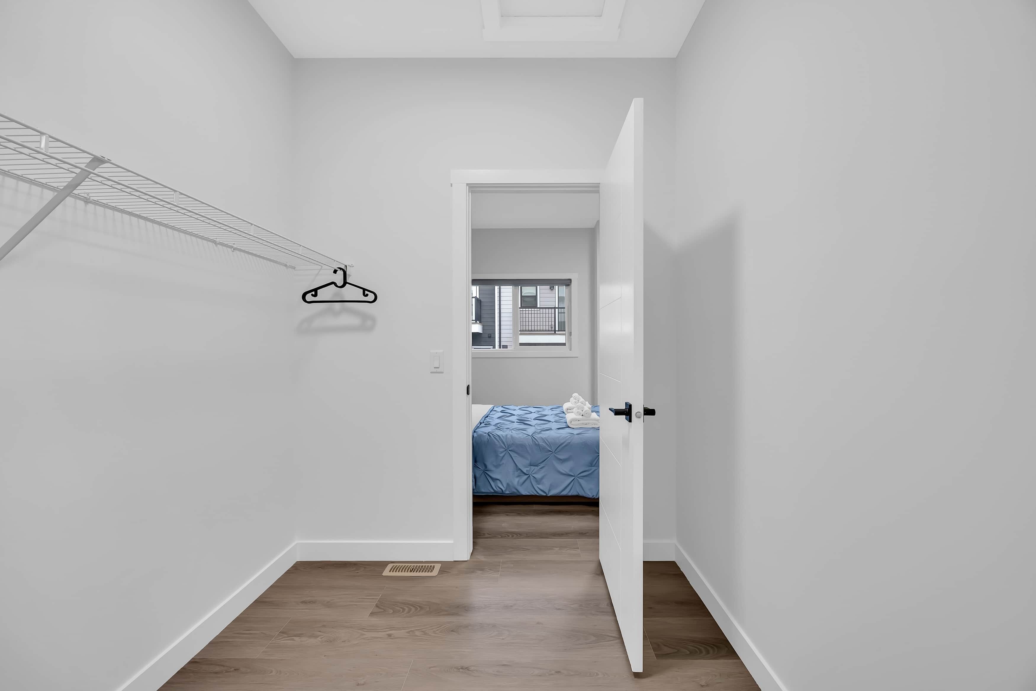 Un couloir étroit avec un porte-vêtements, une porte donnant sur une chambre à coucher où se trouve un lit, et une fenêtre.