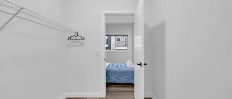Un couloir étroit avec un porte-vêtements, une porte donnant sur une chambre à coucher où se trouve un lit, et une fenêtre.