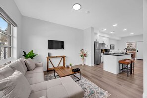 A modern living room with a sofa, coffee table, and a TV. A kitchen with a fridge, stove, and island is visible in the background. - Crescent Heights Oasis (Calgary)