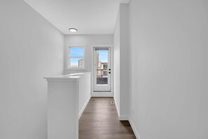 A narrow hallway with white walls and wooden flooring, leading to a door with a view of buildings outside. - Crescent Heights Oasis (Calgary)