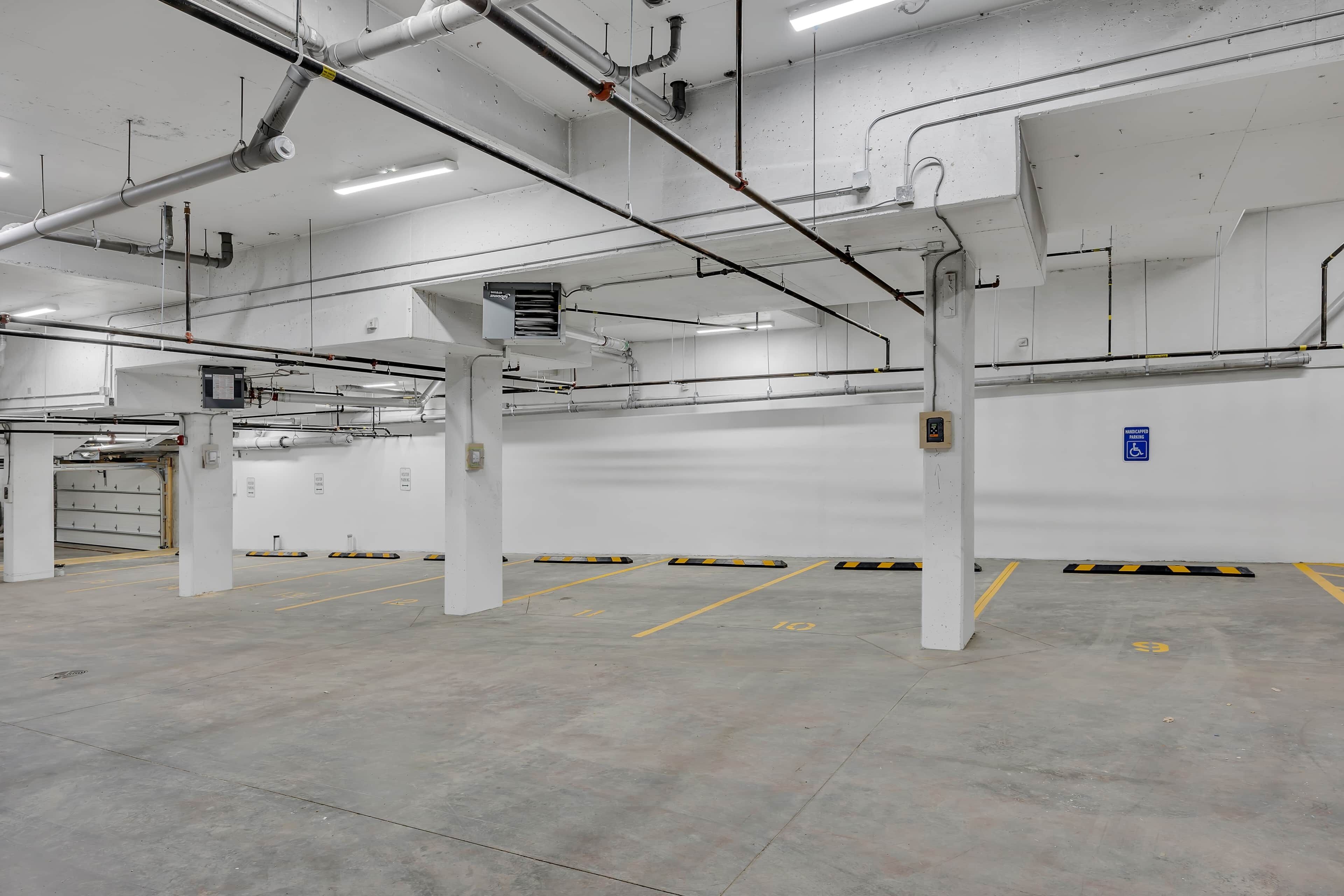 Un estacionamiento amplio y limpio, con paredes blancas, pisos de concreto y elementos estructurales visibles como tuberías y columnas.
