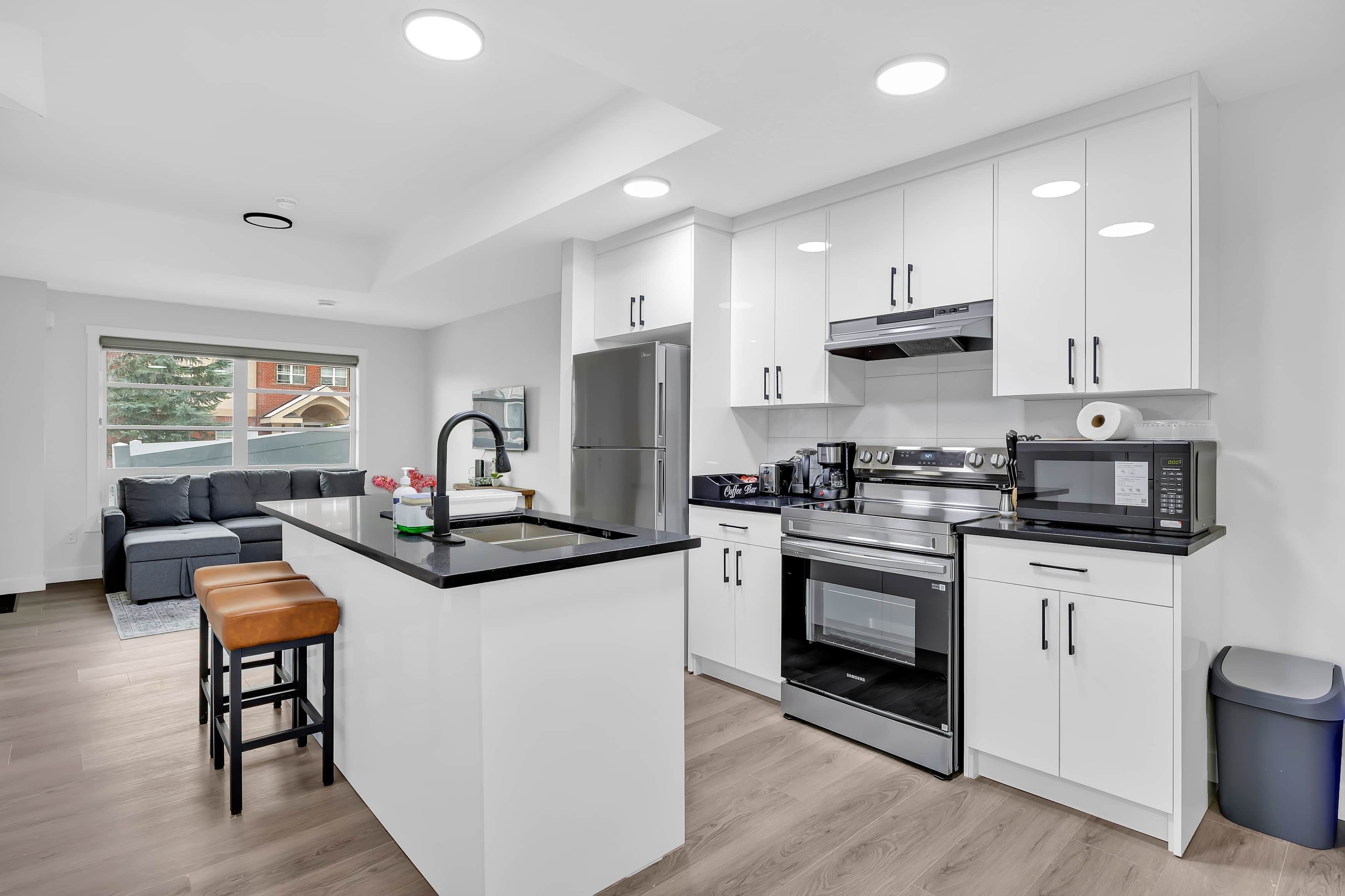 A modern kitchen with white cabinets, stainless steel appliances, a central island, and a grey sofa in the adjacent living area.