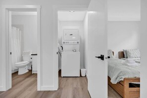 Une chambre à coucher moderne, aux murs blancs, avec un lit en bois, une armoire blanche et une petite salle de bain équipée de toilettes et d’un lave-linge.