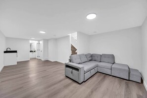 Een moderne woonkamer met een grijze modulaire bank, houten vloeren, witte muren en inbouwspots.