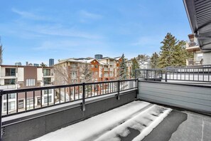 A balcony with a metal railing, a snow-covered ground, and a view of multi-story buildings in the background. - Work away from home! Crescent Heights. (Calgary)