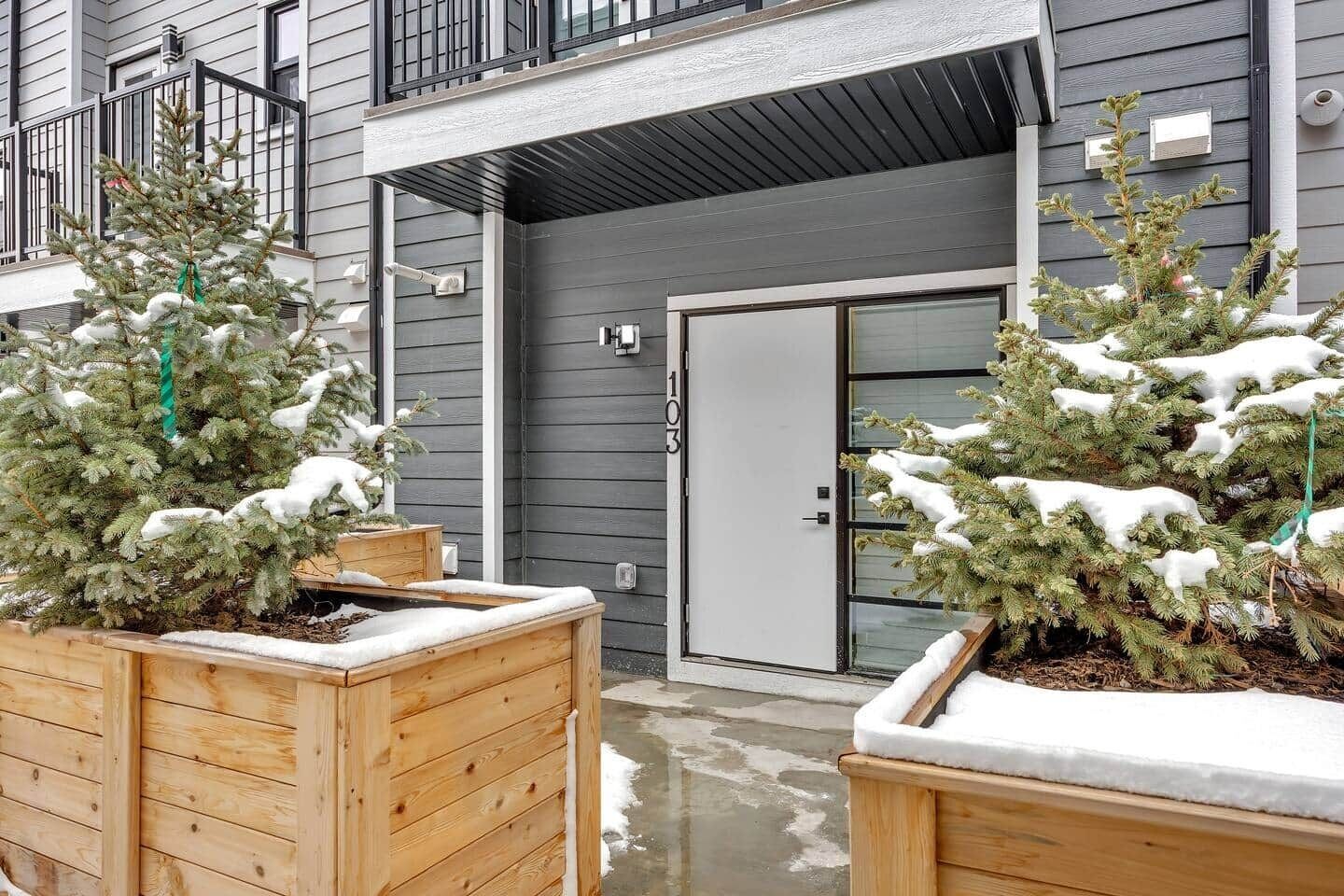 A building with a grey exterior, a balcony, and two wooden planters with snow-covered evergreen trees.