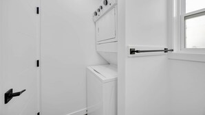 Uma lavanderia com máquina de lavar e secadora, uma porta com puxador e uma janela.