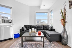 A modern living room with a sectional sofa, a coffee table, a vase, and a microwave in the kitchen area. - Urban Cozy Loft ! 5 min to Downtown (Calgary)