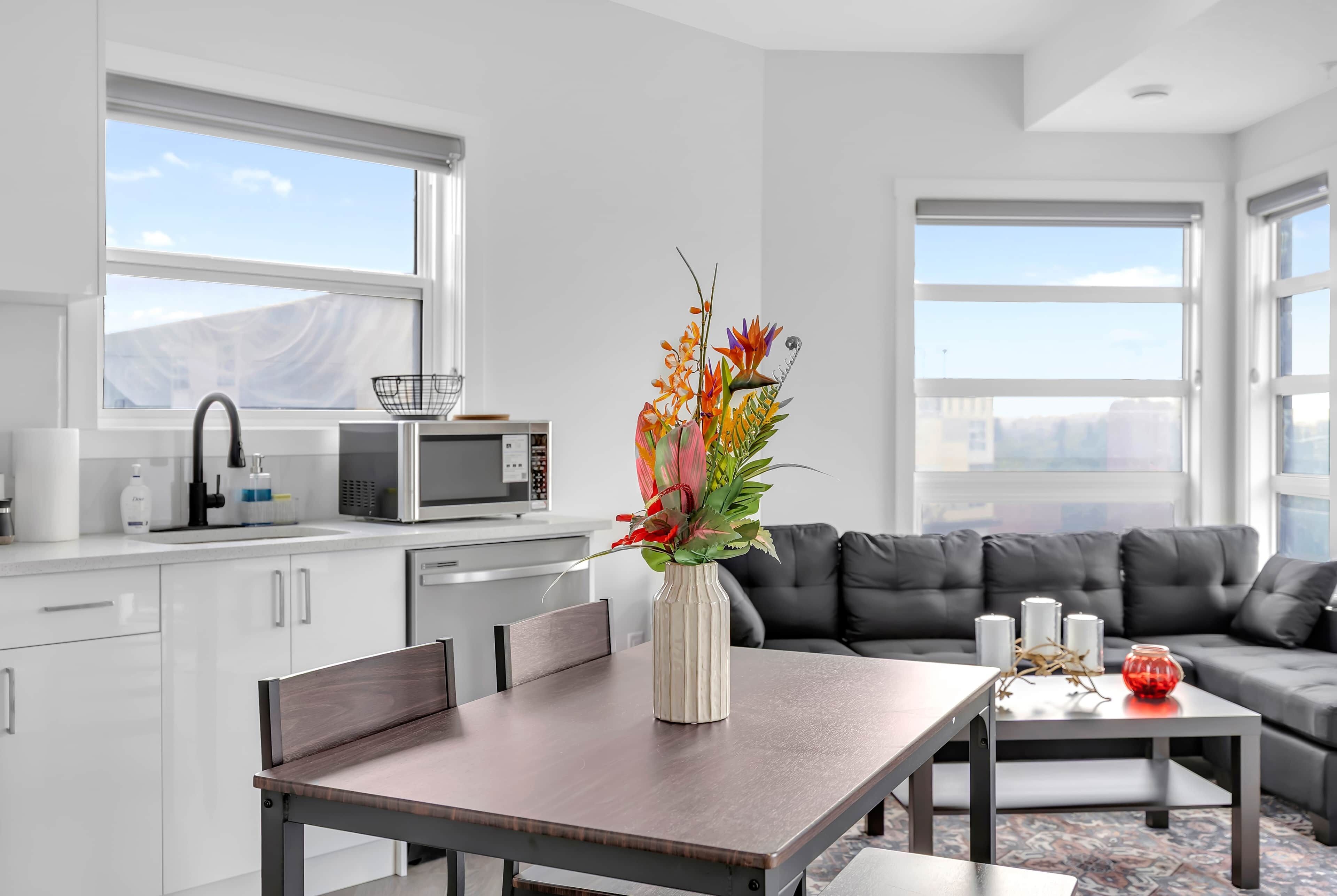 A modern kitchen with white cabinets, a microwave, and a sink. There is a dining table with a vase of flowers. A gray sofa and a coffee table are in the living area.