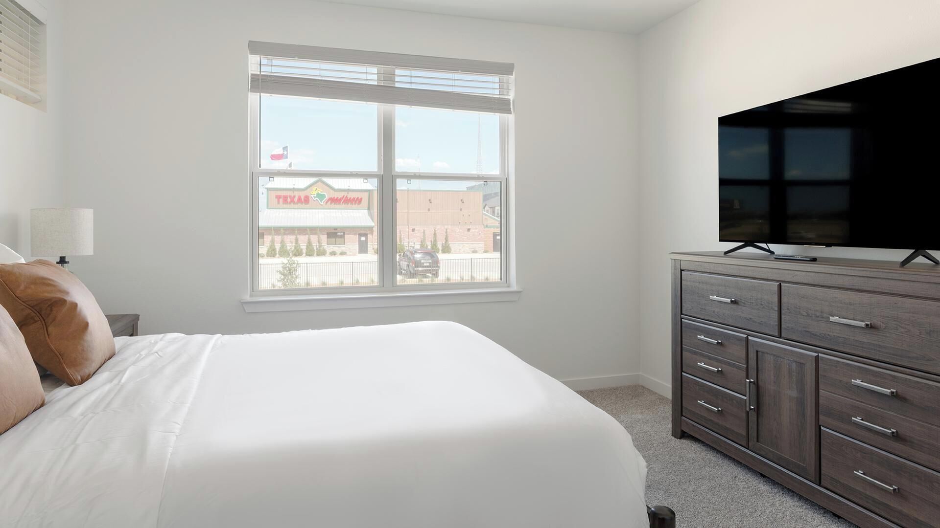 Ein Schlafzimmer mit einem Bett, einer Kommode, einem Fernseher und einem Fenster mit Blick auf ein Gebäude.
