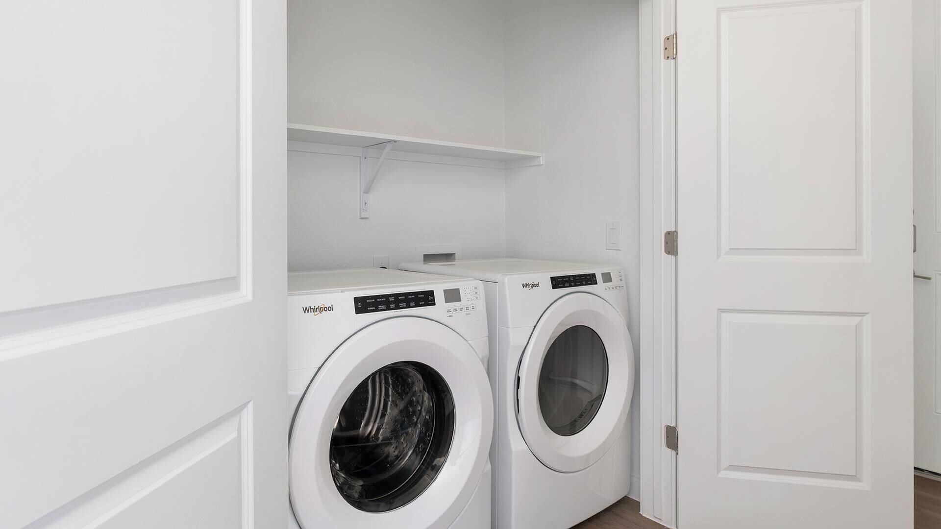 Une buanderie équipée de deux machines à laver Whirlpool blanches, d’une étagère blanche placée au-dessus et d’une porte blanche.