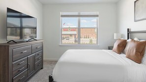 Ein Schlafzimmer mit Bett, Kommode, Fernseher und einem Fenster mit Blick auf ein Gebäude.