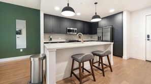 A modern kitchen with dark cabinetry, a white countertop, stainless steel appliances, and two bar stools. - Landing | Spectacular 1BD, Rooftop Lounge, Gym (Denver)