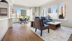 A modern living room with a sofa, armchair, coffee table, and dining area. Large windows offer city views. - Landing | Stylish 2BD, Rooftop Lounge, Gym (Denver)
