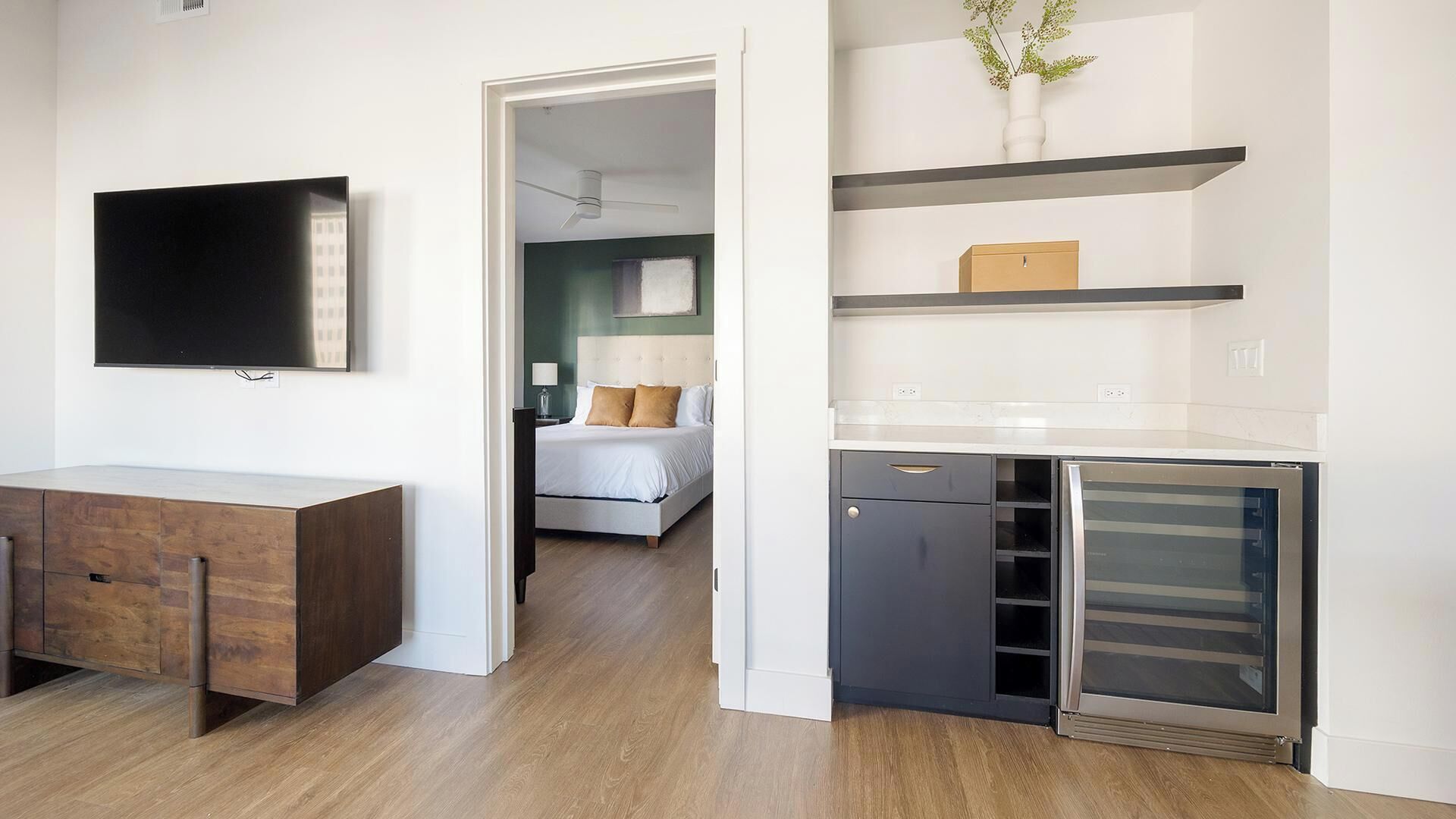 Ett modernt hotellrum med en platt-TV, ett skrivbord i trä, en säng med vita sängkläder, ett sängbord, ett litet bord vid sängen, en lampa, en liten växt, en inbyggd ugn och ett minikylskåp.