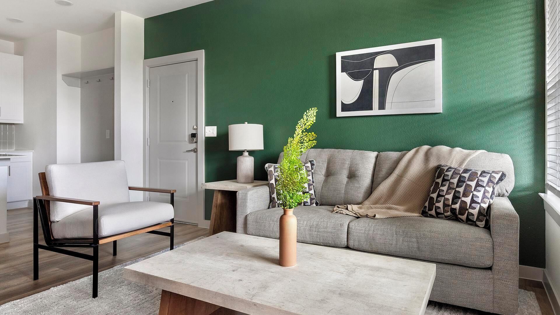 Ein Wohnzimmer mit grauem Sofa, einem weißen Sessel, einem Couchtisch, einer Vase mit Pflanze, einer Lampe und einem gerahmten abstrakten Kunstwerk an der Wand.