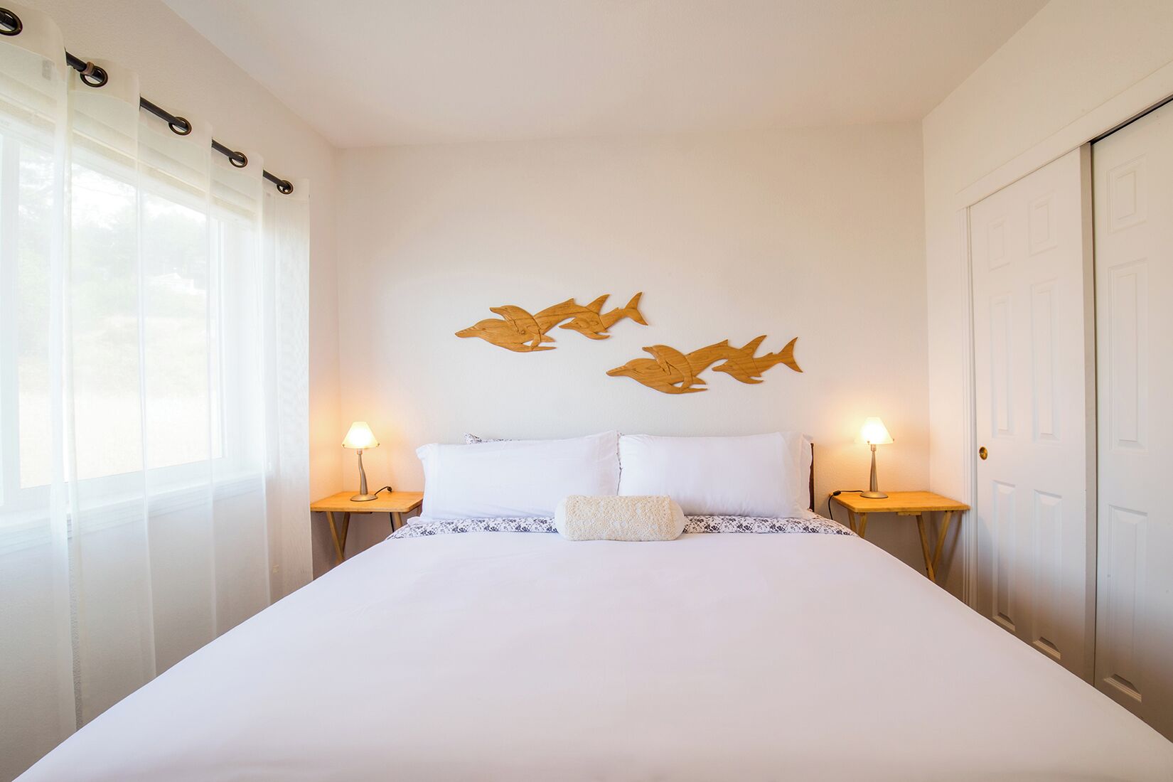 A bedroom with a large bed, two bedside tables with lamps, sheer curtains, and wooden wall art of fish.