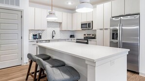 Una cocina moderna con armarios blancos, una isla central con taburetes de bar, electrodomésticos de acero inoxidable e iluminación colgante.