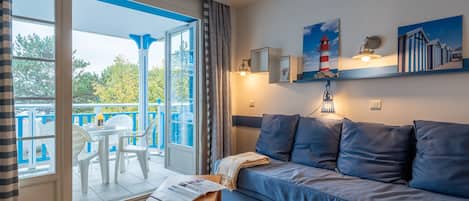 Une pièce avec un canapé bleu, une table en bois et un balcon avec des chaises blanches.
