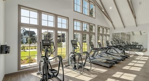 Un gimnasio con cintas de correr y elípticas múltiples, amplias ventanas con vista a una zona residencial, y un techo alto con vigas de madera.
