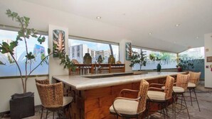 Una zona de bar con mostrador de madera y sillas de mimbre, ventanas amplias con vistas a la ciudad y plantas de interior.