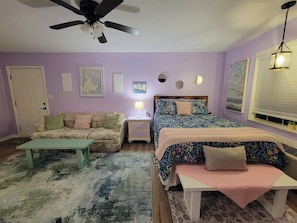 A bedroom with a bed, a sofa, a nightstand, a lamp, a ceiling fan, and a window with blinds.