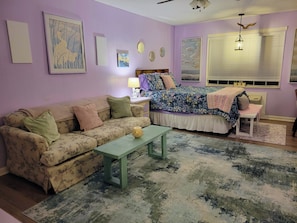 A room with a floral-patterned sofa, a bed with a floral bedspread, a small table, and a window with blinds.